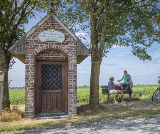 Twee mannen rusten op een bankje naast een kapelletje in een groen veld, met fietsen geparkeerd onder een boom. (vergroot foto)